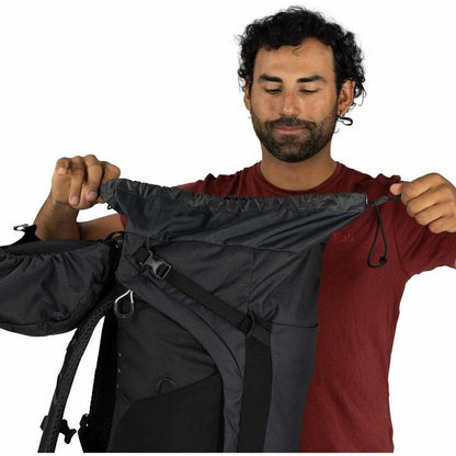 A man in a red shirt holds open the top of the Moss Green Osprey Kestrel LT 65 backpack against a white background.