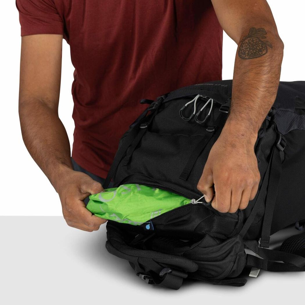 A person in a red shirt opens the front pocket of the black Osprey Kestrel LT 65 backpack, revealing a bright green item inside.