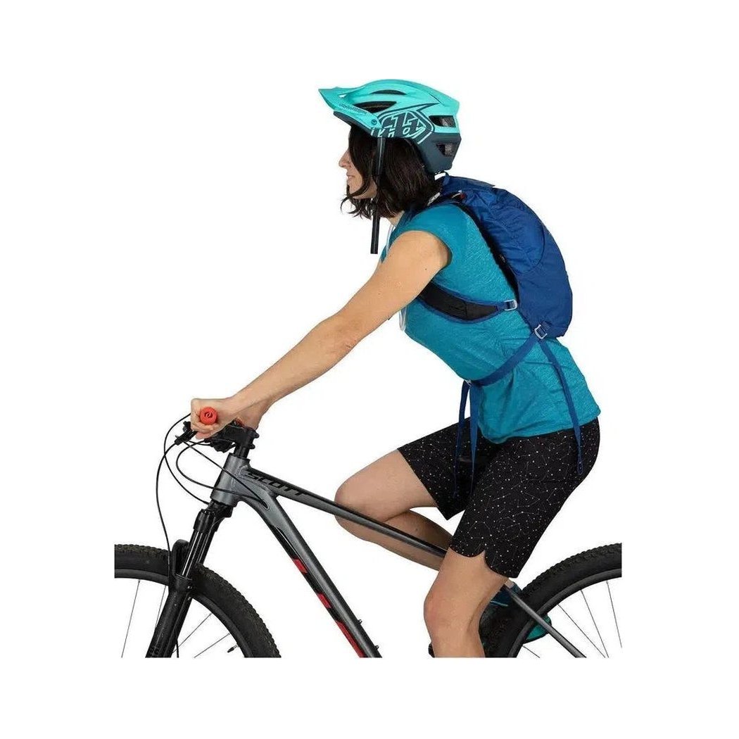 Side view of the Osprey Kitsuma 7 in SpaceTravelGrey showing the blue backpack and teal helmet on a model riding a bike against a white background.