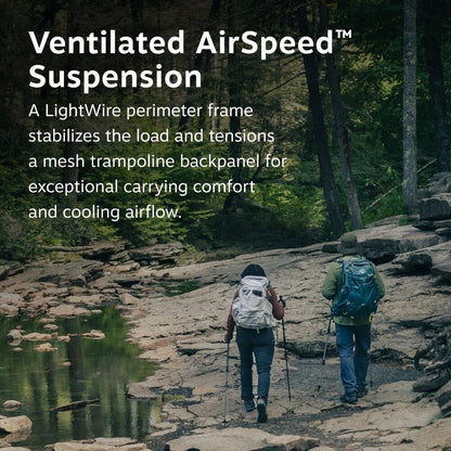 Two hikers on a rocky trail wear Osprey Manta 24 Hiking Hydration Backpacks in Dark Charcoal, with a forest and stream in the background.