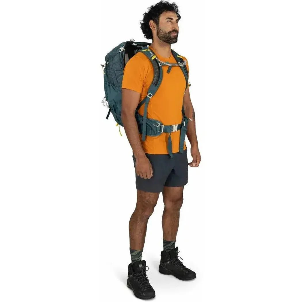 A man in an orange shirt and dark shorts wears the Osprey Manta 34 Hiking Hydration Backpack in Dark Charcoal, shown from the side against a white background.