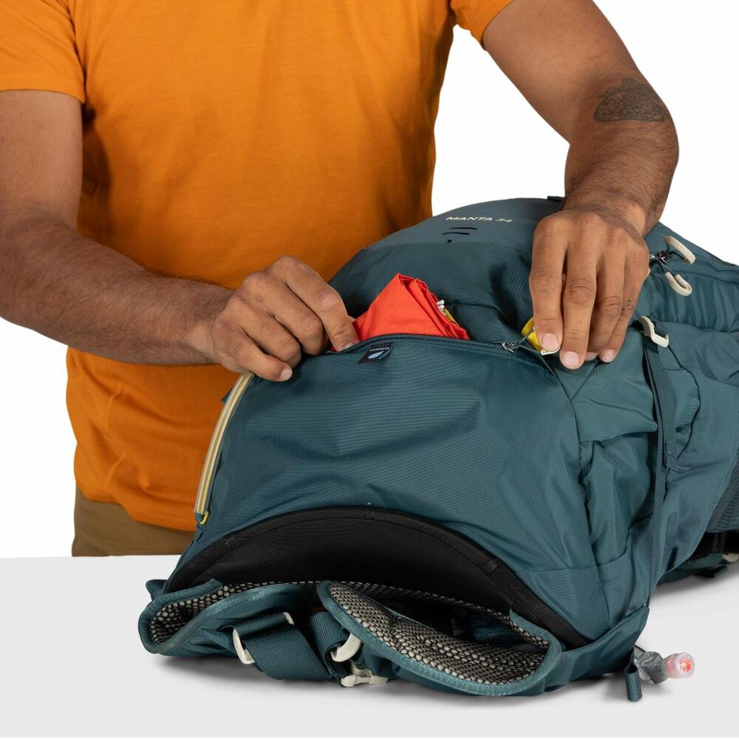 A person in an orange shirt zips the Osprey Manta 34 Hiking Hydration Backpack with a red item visible in the top pocket, against a white background.