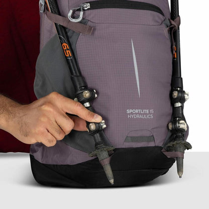 A hand adjusts the trekking pole attachment on the Graphite Purple Osprey Sportlite 15 Hydraulics backpack against a white background.