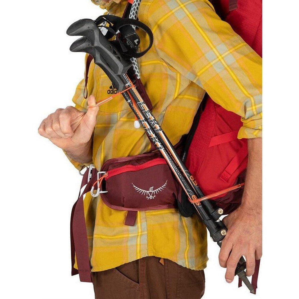 A person in a yellow plaid shirt wears the Osprey Stratos 24 backpack with a red pack and maroon waist pack visible.