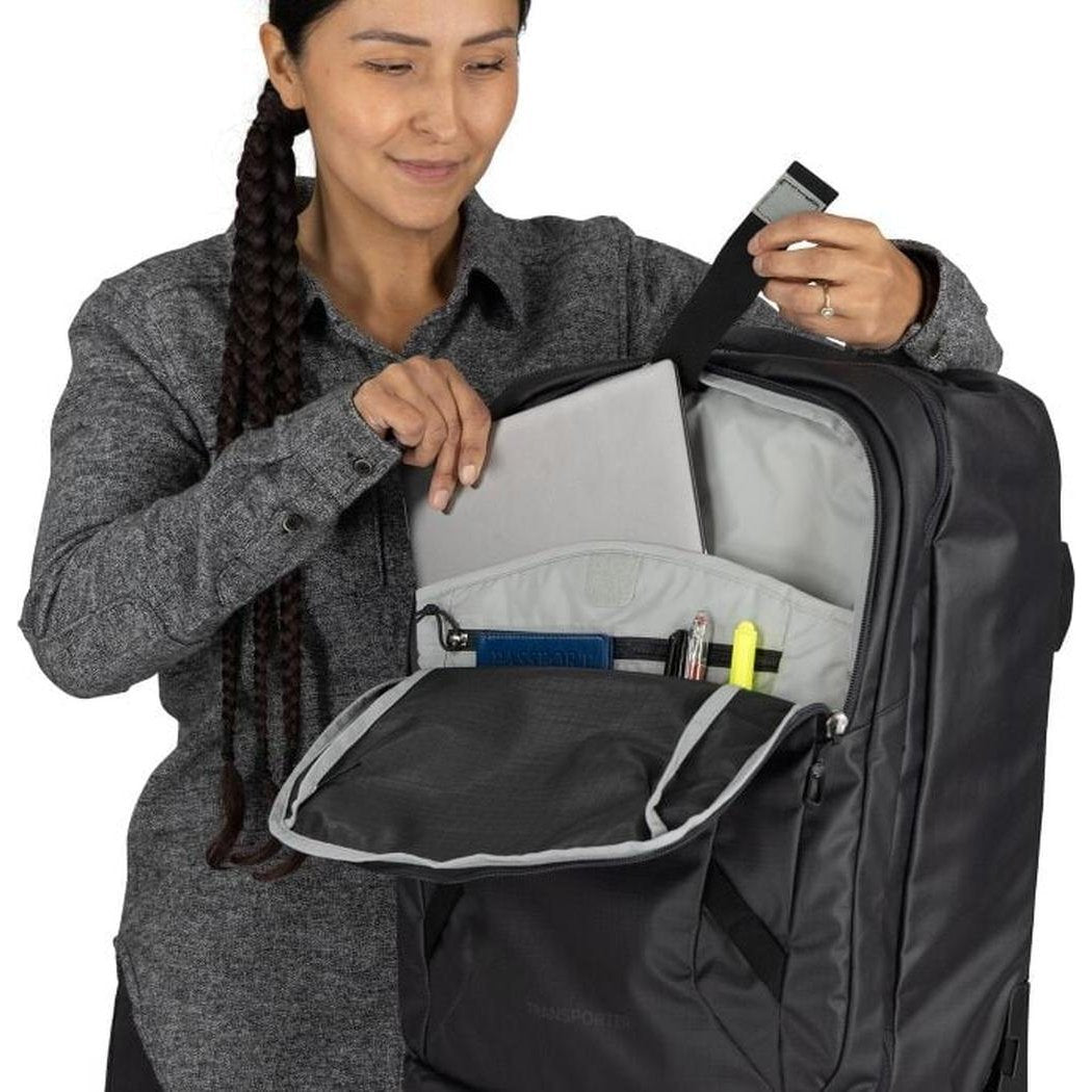 A woman wearing a gray shirt holds the Osprey Transporter Wheeled Carry On in Raven Black, showing its open front pocket with pens and a laptop inside against a white background.
