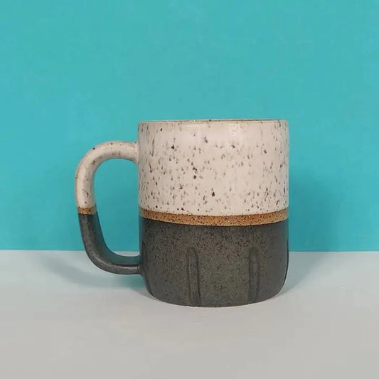 A side view of the Osso Ceramics Black and White Cookie Mug showing its speckled top and charcoal bottom against a turquoise background.