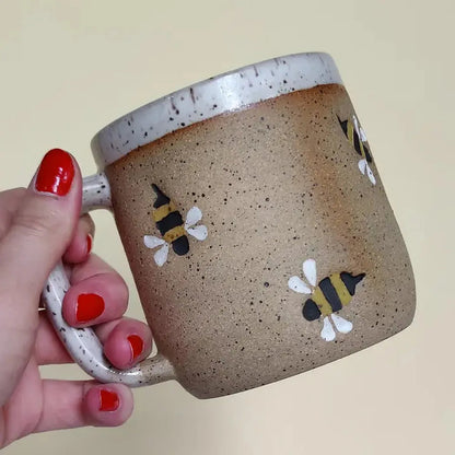 A hand with red nail polish holds the Osso Ceramics Bumble Bee Stamped Mug against a light background.