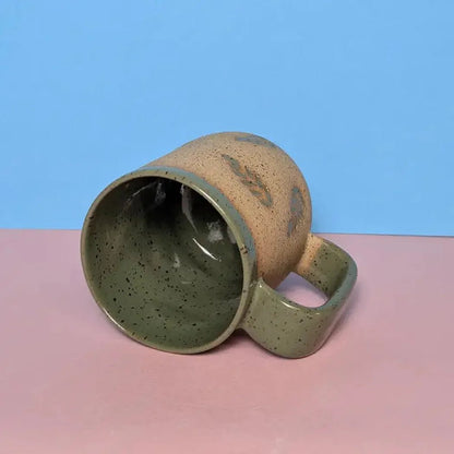 A side view of the Osso Ceramics Fern Stamped Mug showing its speckled interior and handle against a blue and pink background.