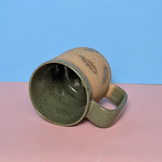 A side view of the Osso Ceramics Fern Stamped Mug showing its speckled interior and handle against a blue and pink background.