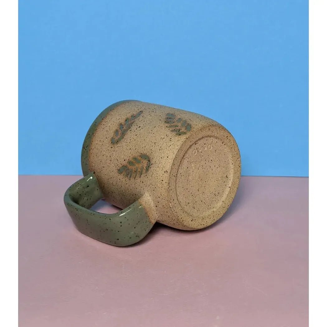 A side view of the Osso Ceramics Fern Stamped Mug with a green handle, lying on a pink surface against a blue background.