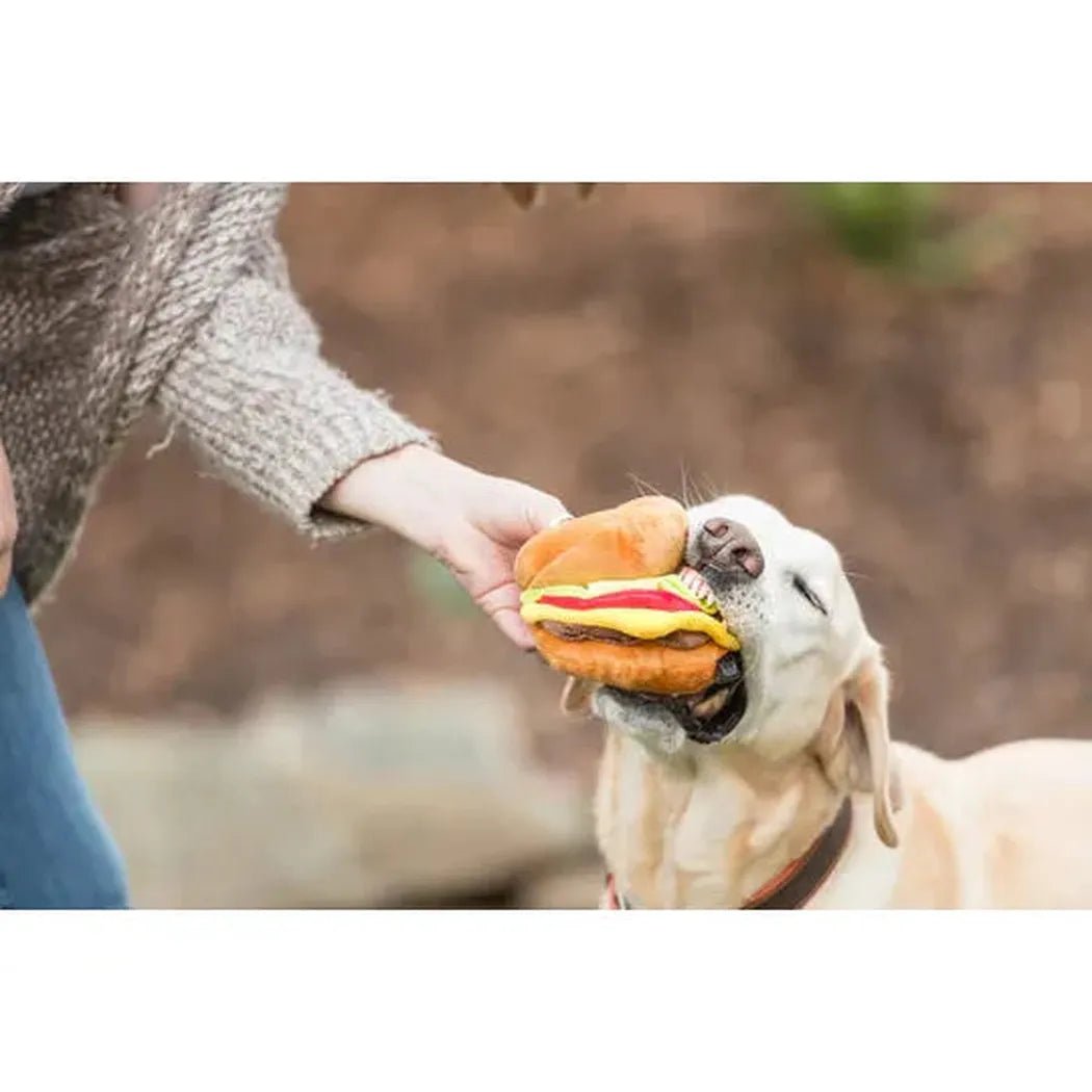 A person offers a P.L.A.Y American Classic Barky Burger toy to a happy yellow dog outdoors.