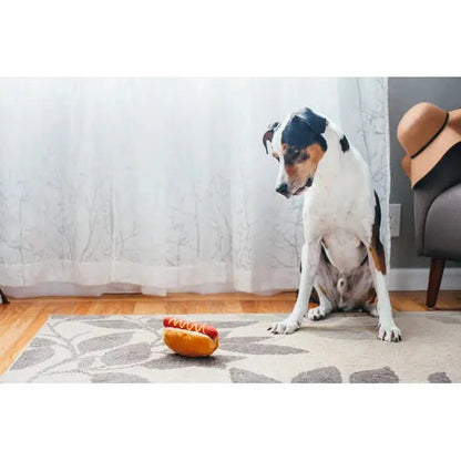 A dog sits on a rug looking at a P.L.A.Y American Classic Hot Diggy Dog toy in front of it.