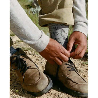 A person tying the laces of brown hiking boots while wearing light grey Paka Apparel Essential Sebastian Crew Socks with a patterned design, on a gravel path.