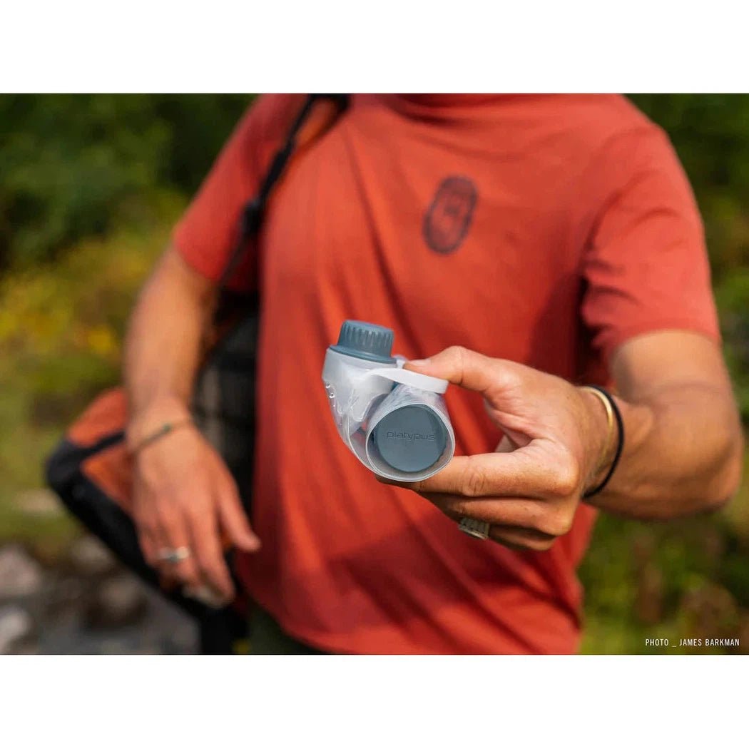 A person in a red shirt holds the Platypus 1L Quickdraw hydration bottle, showing its gray and blue design against a green outdoor background.