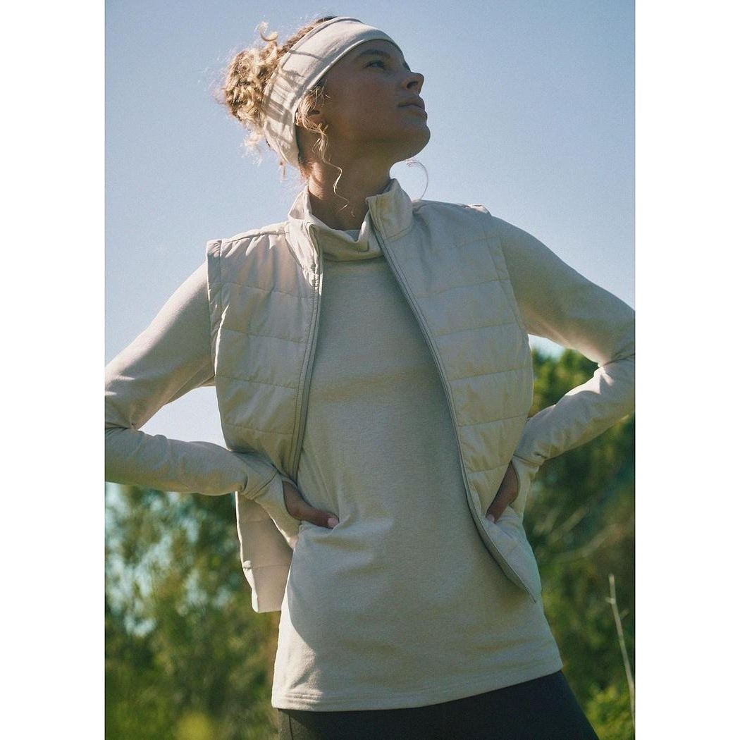 A woman wearing the Prana Women's Ice Flow Long Sleeve in Almond Heather stands outdoors with her hands on her hips against a blue sky and green trees.