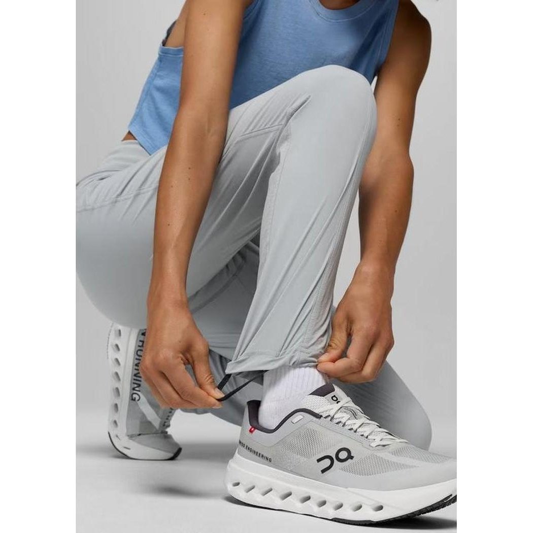 A woman crouching adjusts her grey blue Prana Women's Koen Mid Rise Daily Pant while wearing white On running shoes against a plain background.
