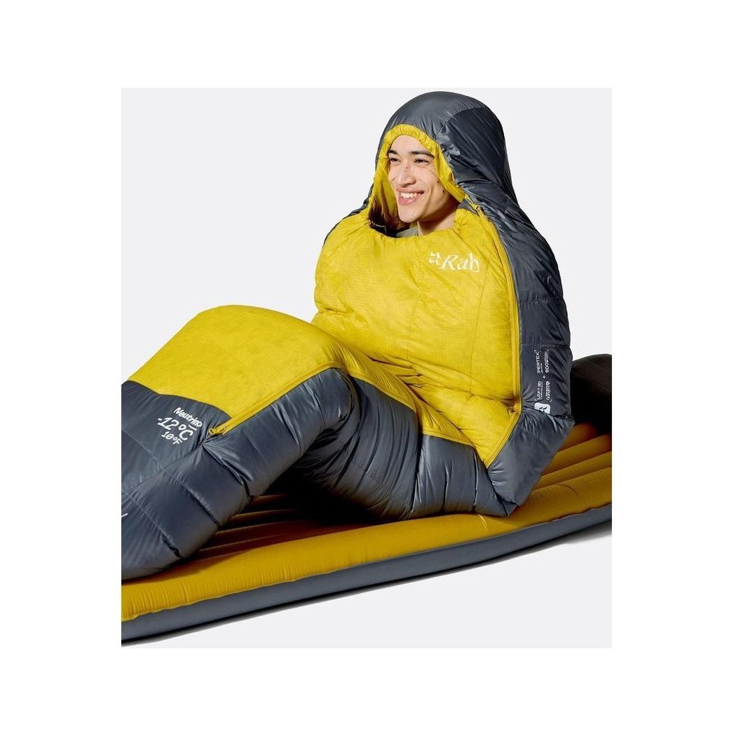 A model wearing the Rab Neutrino 0C / 32F sleeping bag in Graphene/Dark Pollen sits on a yellow and gray mat.