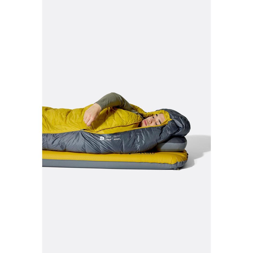 A person wearing the Rab Neutrino 0C / 32F Graphene/Dark Pollen sleeping bag lies on a yellow and gray air mattress against a white background.