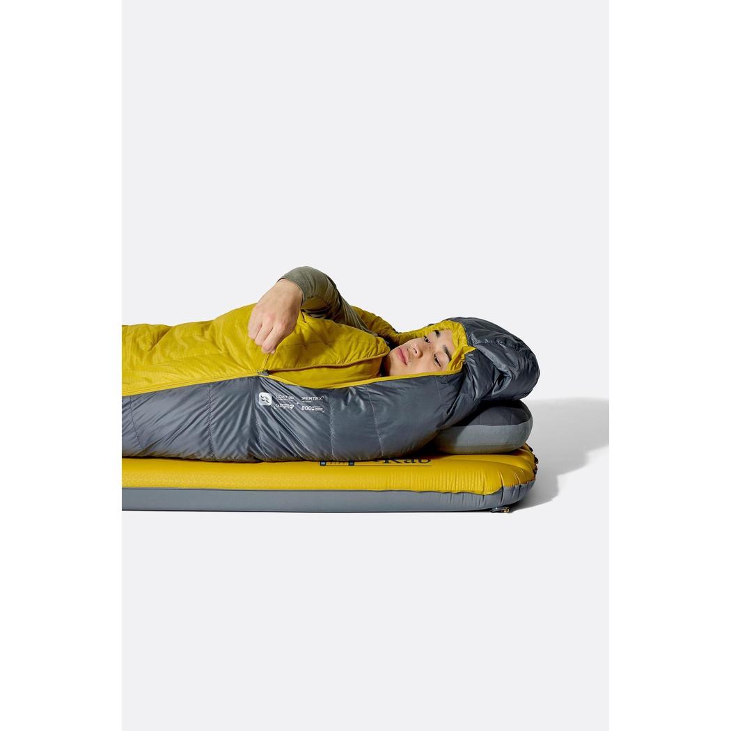 A person wearing the Rab Neutrino 0C / 32F Graphene/Dark Pollen sleeping bag lies on a yellow and gray air mattress against a white background.