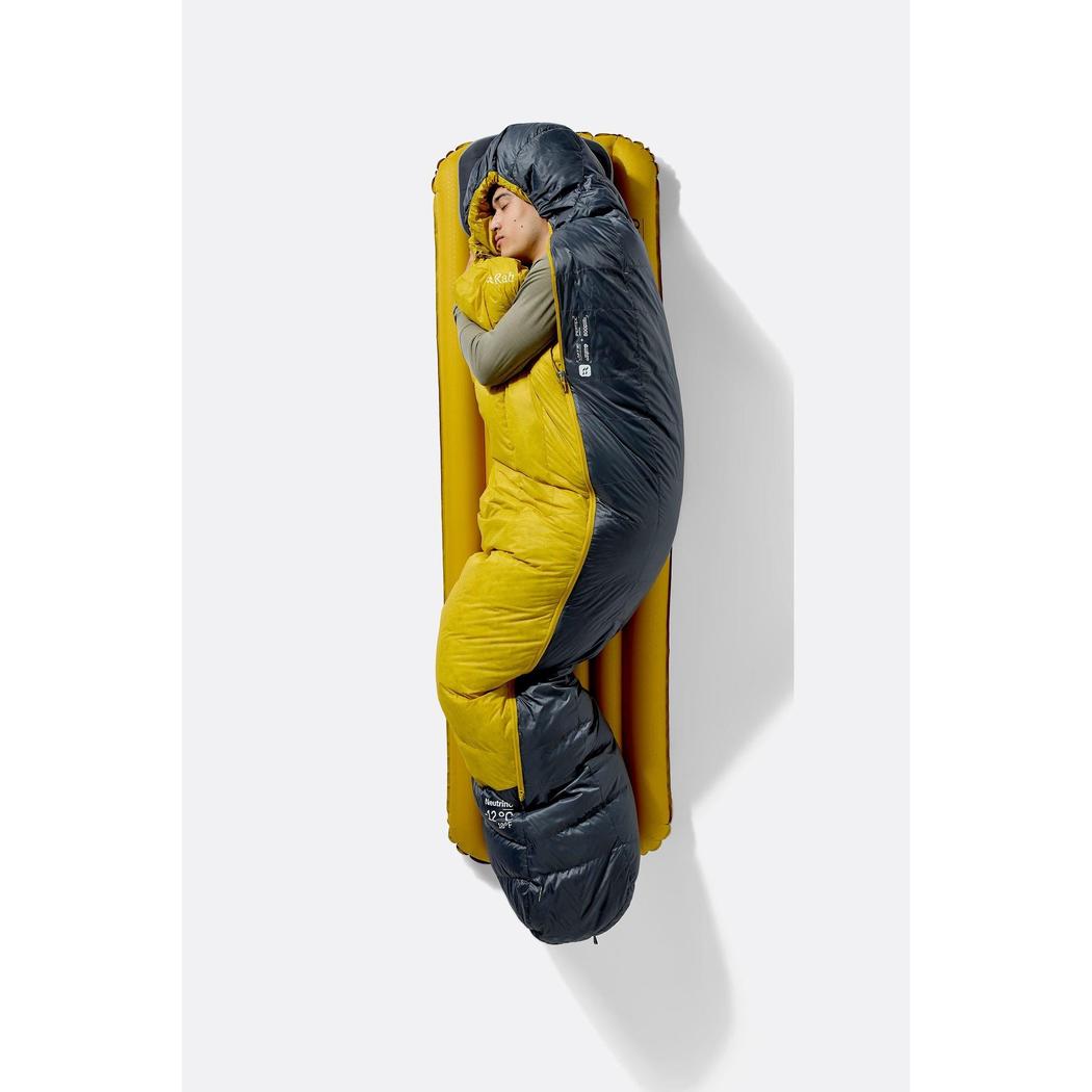 A person wearing the Rab Neutrino 0C / 32F sleeping bag in Graphene/Dark Pollen lies on a yellow sleeping pad against a white background.