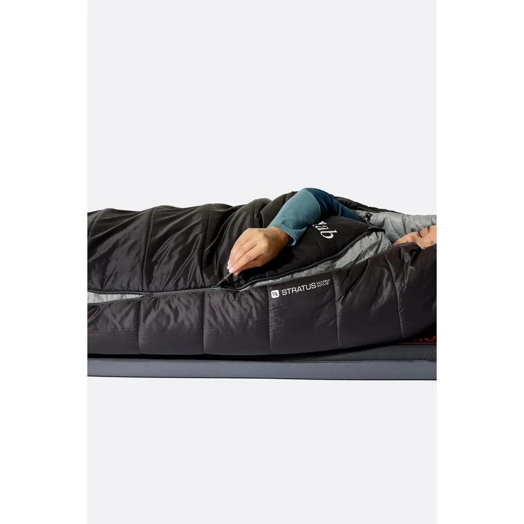 A woman wearing a teal long-sleeve lies in a dark green Rab Radeon sleeping bag with a left zip, shown from a top-down angle on a gray surface.