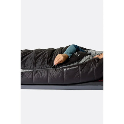 A woman wearing a teal long-sleeve lies in a dark green Rab Radeon sleeping bag with a left zip, shown from a top-down angle on a gray surface.