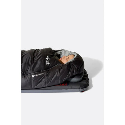 A woman lies in a dark green Rab sleeping bag on a gray mat against a white background.