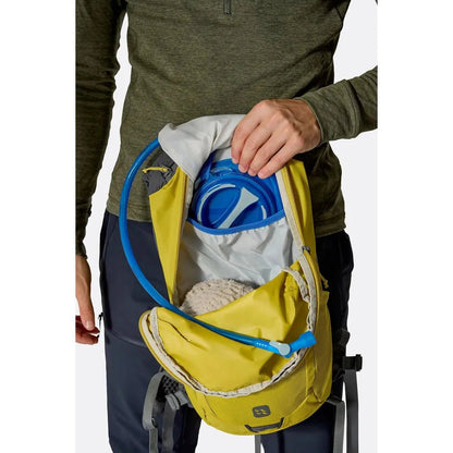 A person wearing a green top holds open the yellow Rab Tygen 12 daypack, showing its blue hydration reservoir and white interior against a white background.