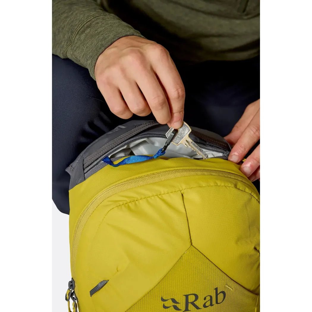 A person zips the yellow Rab Tygen 12 daypack, showing its top compartment and logo.