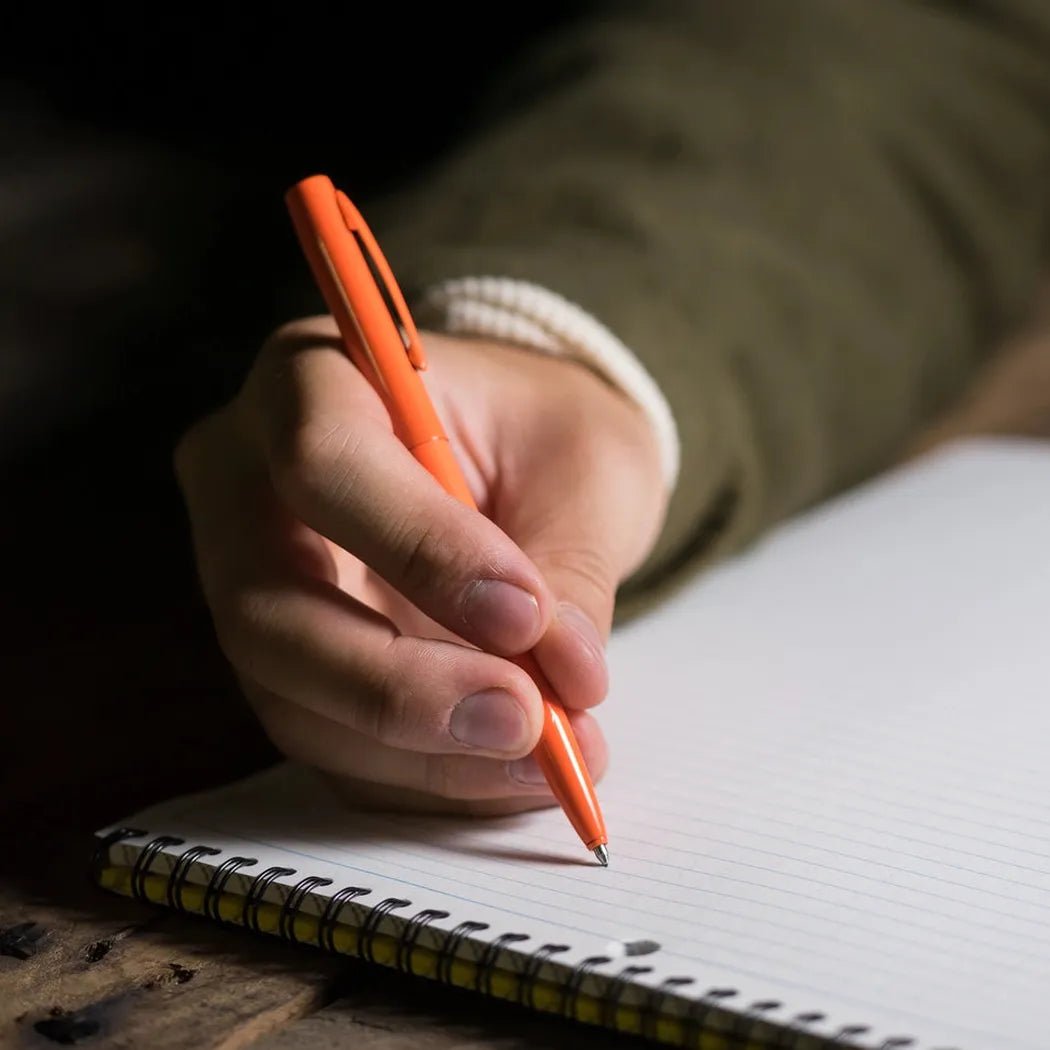 A hand holds the Rite in the Rain All-Weather Metal Pen in black over a spiral notebook on a wooden surface.
