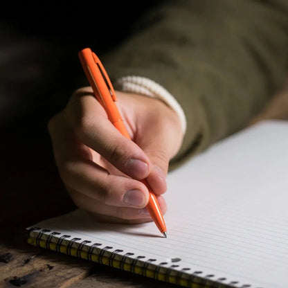 A hand holds the Rite in the Rain All-Weather Metal Pen in black over a spiral notebook on a wooden surface.