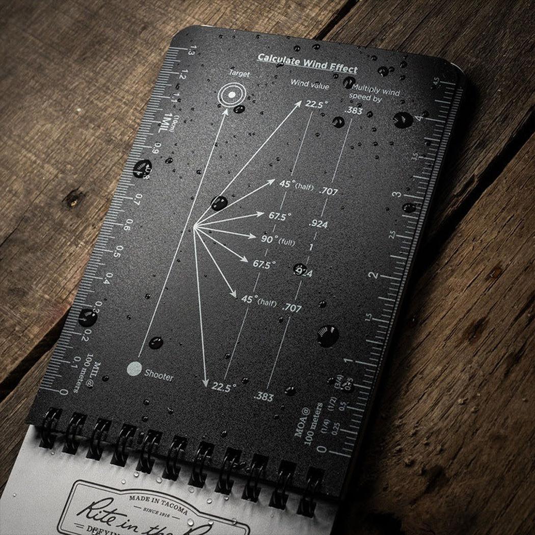 A black Rite in the Rain Dope Logbook - Top Spiral 4x6 lies on a wooden surface with water droplets and wind effect calculations visible.