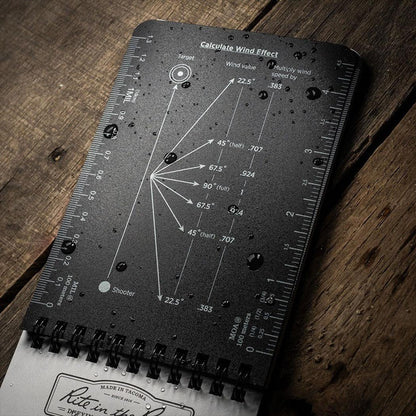 A black Rite in the Rain Dope Logbook - Top Spiral 4x6 lies on a wooden surface with water droplets and wind effect calculations visible.