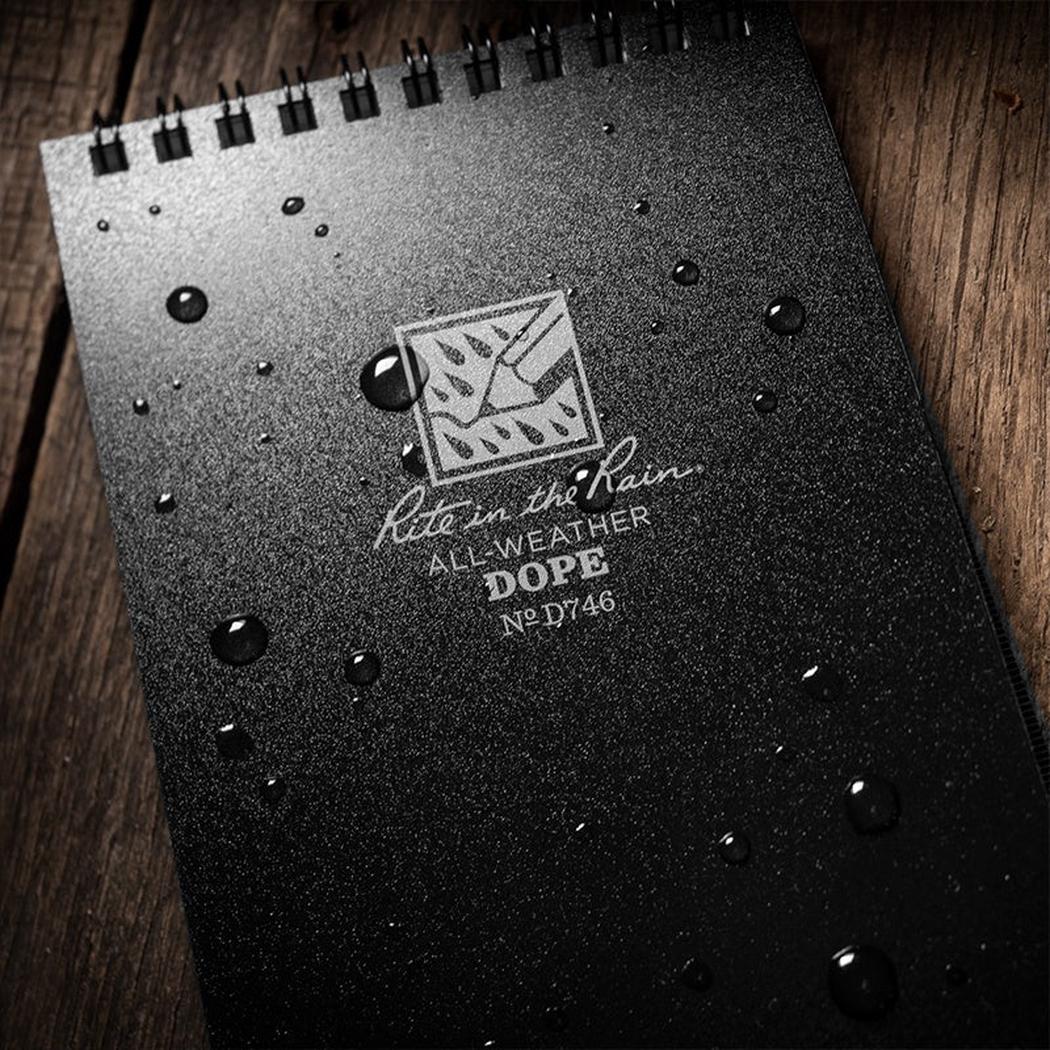 A top-down view of the black Rite in the Rain Dope Logbook with water droplets on its cover against a wooden background.