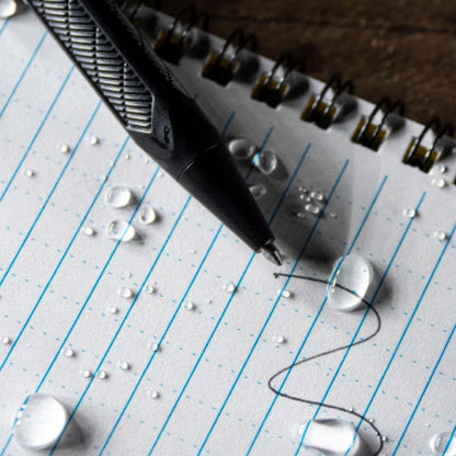 A close-up of the Black Rite in the Rain Mini Bolt-Action Pen on a lined notebook with water droplets.
