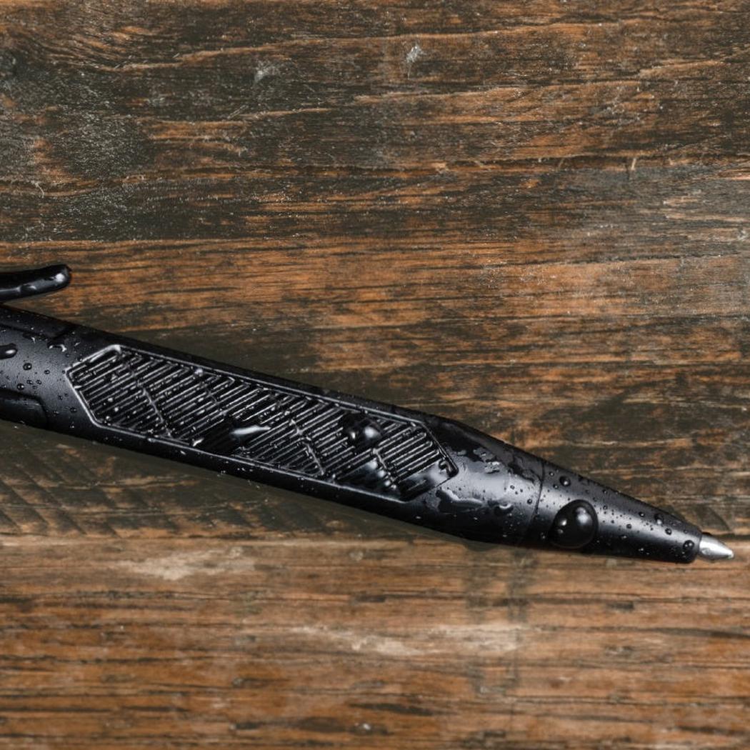 A black Rite in the Rain Mini Bolt-Action Pen lies on a weathered wooden surface with water droplets visible on its body.