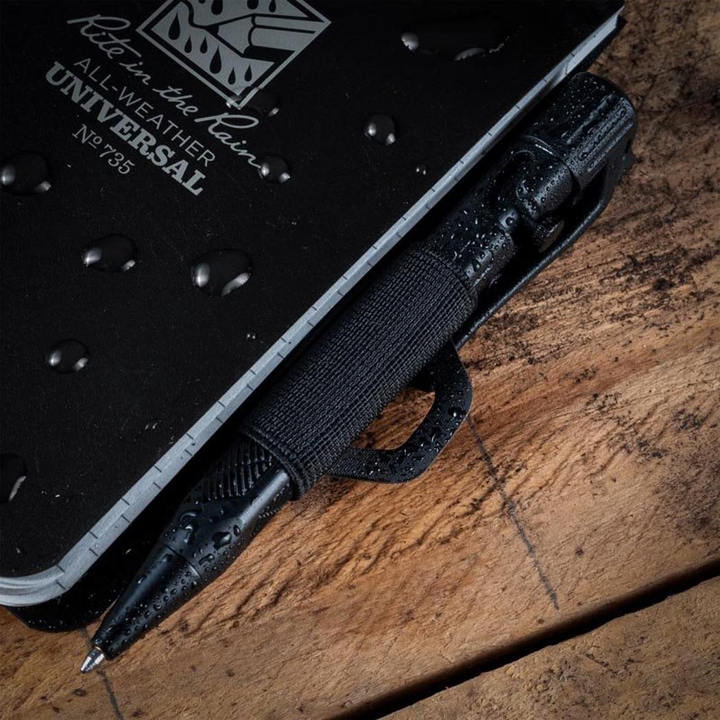 A close-up of the Rite in the Rain Monsoon Pen Holster in Dark Green, with a black notebook and pen resting on a wooden surface.