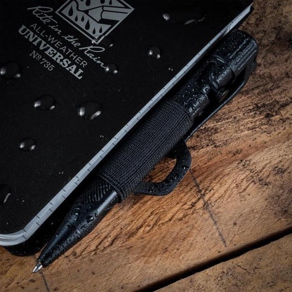 A close-up of the Rite in the Rain Monsoon Pen Holster in Dark Green, with a black notebook and pen resting on a wooden surface.