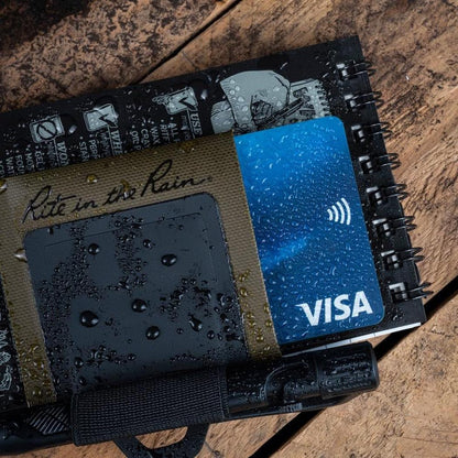 A top-down view of the Rite in the Rain Monsoon Pen Holster in Dark Green holding a Visa card on a wooden surface.