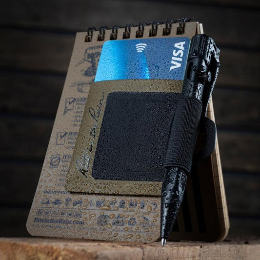 A close-up of the Rite in the Rain Monsoon Pen Holster in Dark Green, showing the notebook and pen with water droplets, against a dark wooden background.