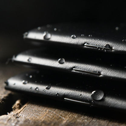Three black Rite in the Rain notebooks stacked on a wooden surface with water droplets.