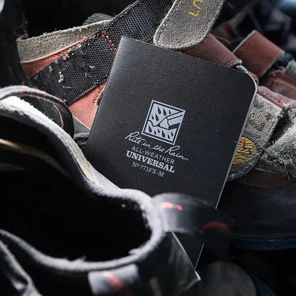 A black Rite in the Rain notebook rests among rugged hiking boots, showing its all-weather design.