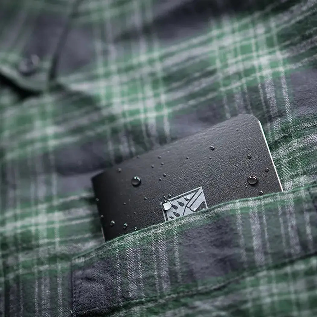 A black Rite in the Rain notebook sits in the pocket of a green plaid shirt, showing water droplets on its surface.