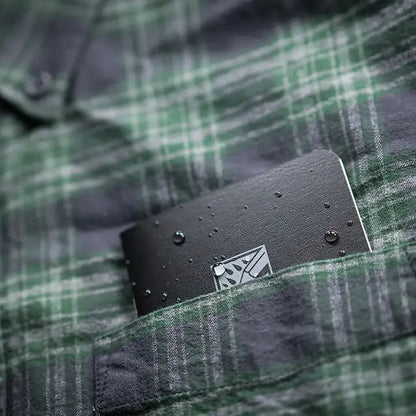 A black Rite in the Rain notebook sits in the pocket of a green plaid shirt, showing water droplets on its surface.