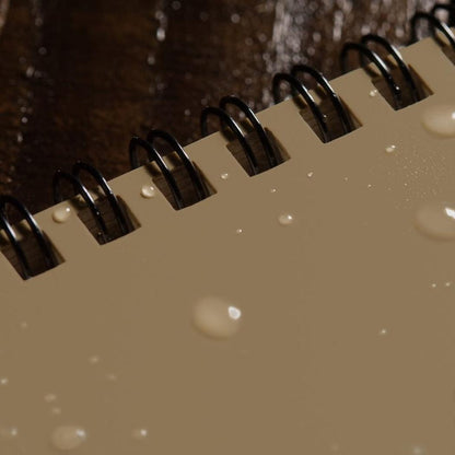 A close-up of the Rite in the Rain Side Spiral Notebook 4 5/8 x 7 showing the black spiral binding and water droplets on the cover.