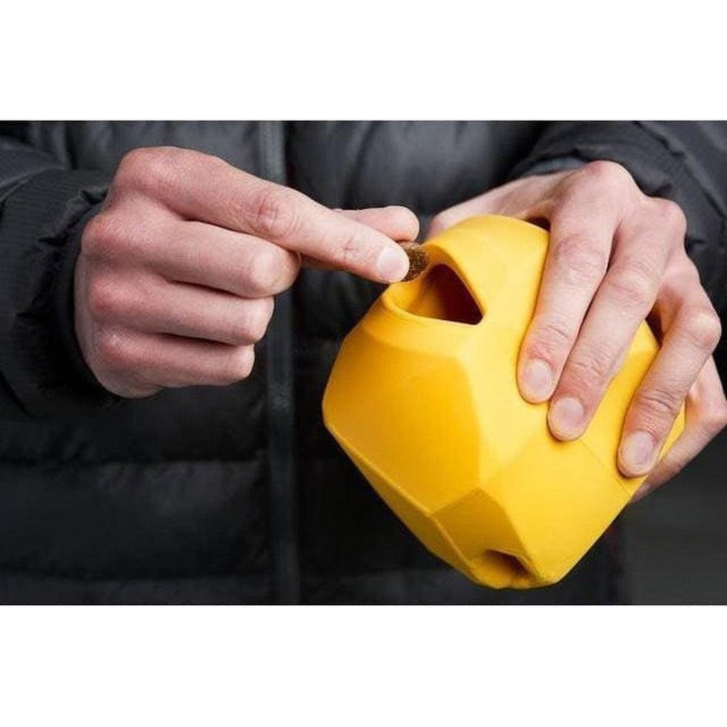 A person in a charcoal jacket holds the Dandelion Yellow Ruffwear Gnawt-a-rock toy, inserting a coin into its triangular opening.