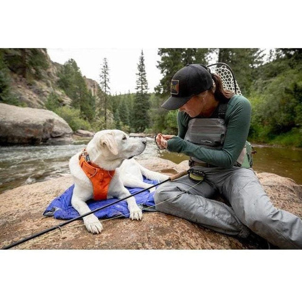 A woman sits on a rock by a river with her white dog wearing an orange vest, both on a Huckleberry Blue Ruffwear Highlands Dog Sleeping Bag.