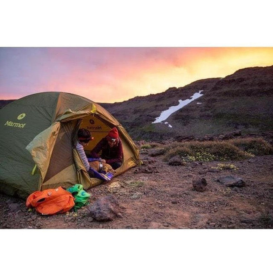 A person sits in a Marmot tent at sunset with an orange and green bag on the ground, and a Huckleberry Blue Ruffwear Highlands Dog Sleeping Bag visible inside.
