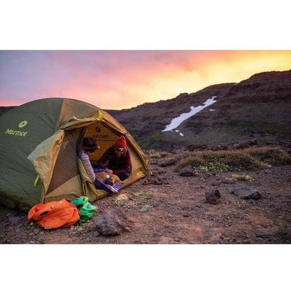 A person sits in a Marmot tent at sunset with an orange and green bag on the ground, and a Huckleberry Blue Ruffwear Highlands Dog Sleeping Bag visible inside.