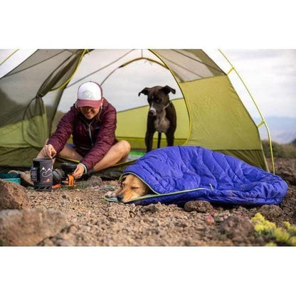 A person prepares coffee outside a tent while a dog sleeps in the Huckleberry Blue Ruffwear Highlands Dog Sleeping Bag.